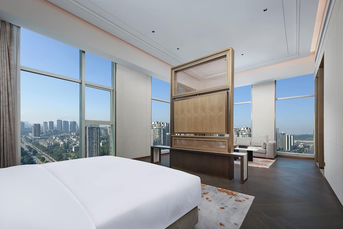 A modern hotel room with a large bed, a seating area with a sofa and armchairs, a TV, and a city view through large windows.