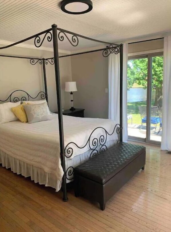 A four-poster bed with a bench, a nightstand with a lamp, and a view of the outdoors through a glass door. - Waterfront Lake House on Wilson Lake (Florence)