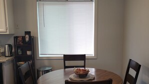 Comedor con una mesa redonda, cuatro sillas, cafetera y ventana con persianas.