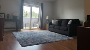 Una sala de estar con un sofá gris oscuro, un tapete estampado azul, piso de madera, un ventanal corredero con cortinas, una mesita auxiliar con una lámpara y un cuadro en un mueble de madera.