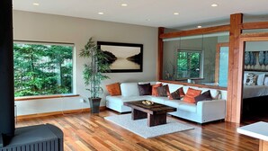 A modern living room with a white sofa, wooden flooring, a large window with a view of trees, and a mirror reflecting the room. - Points West Oceanfront Resort 1 Bed Ocean Point Suite B (Sooke)