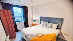 Habitación de hotel con una cama grande, una mesita de noche con lámpara, una ventana con cortinas y vistas a la ciudad.