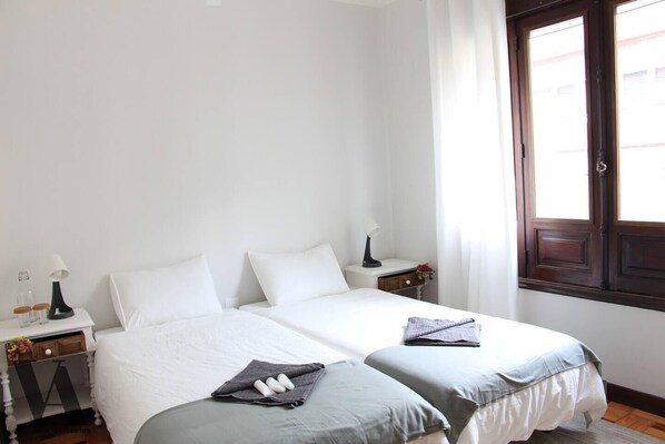 A hotel room with two single beds, a nightstand with a lamp, a small table with a vase, and a window with curtains.