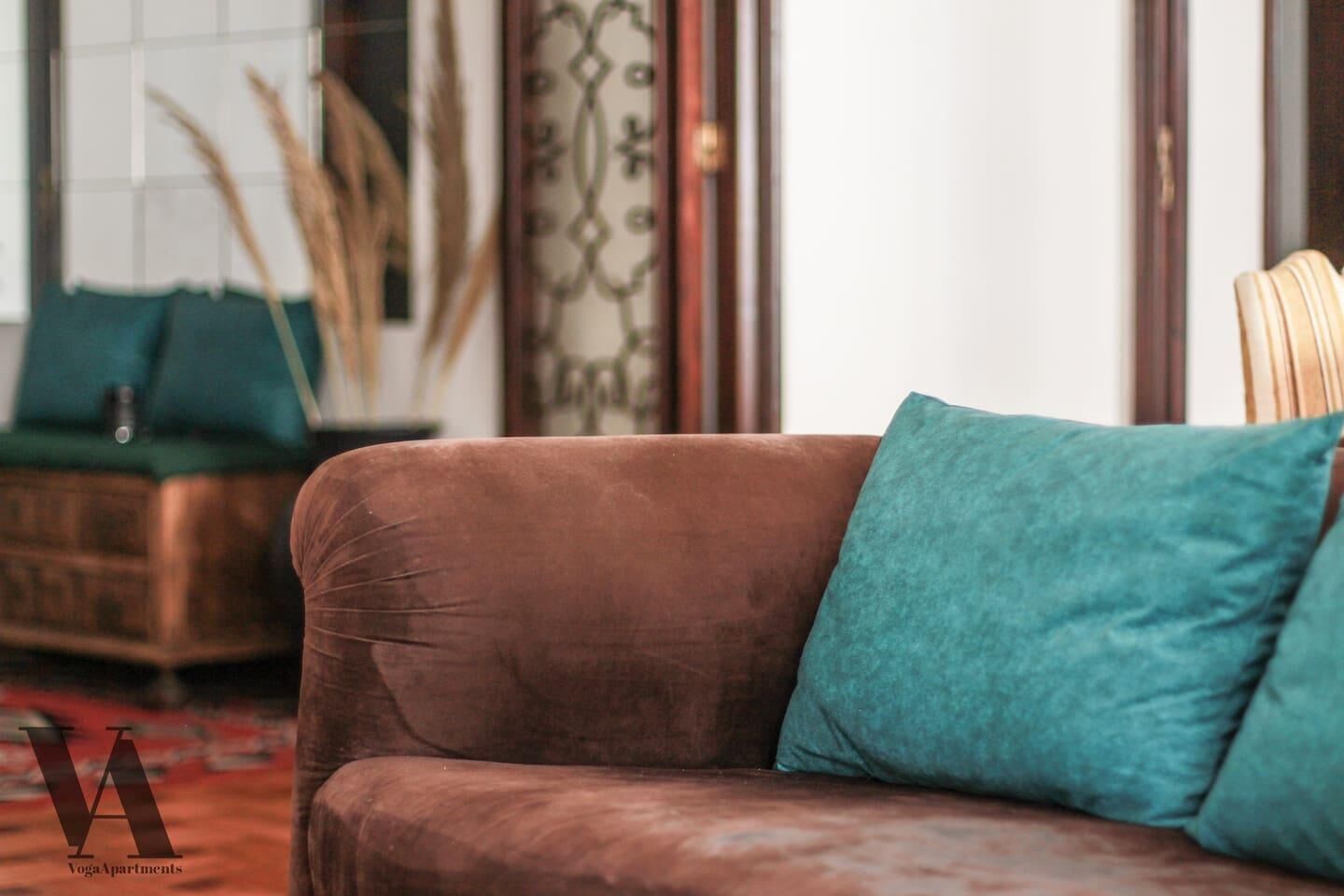 Ein braunes Sofa mit türkisblauen Kissen in einem Raum mit einem gemusterten Vorhang und einem grünen Kissen auf einem passenden Sofa.