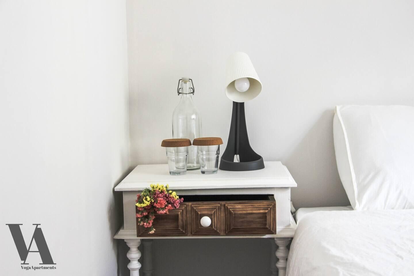 A white bedside table with a lamp, two glass bottles with cork lids, and a small wooden drawer.