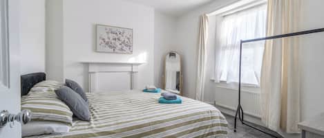 Una habitación de hotel con cama, espejo, ventana con cortinas, radiador y un cuadro colgado en la pared.