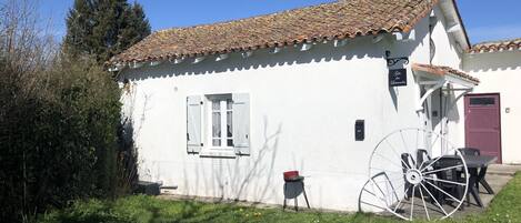 A white building with a tiled roof, a window with shutters, and a large wheelbarrow.