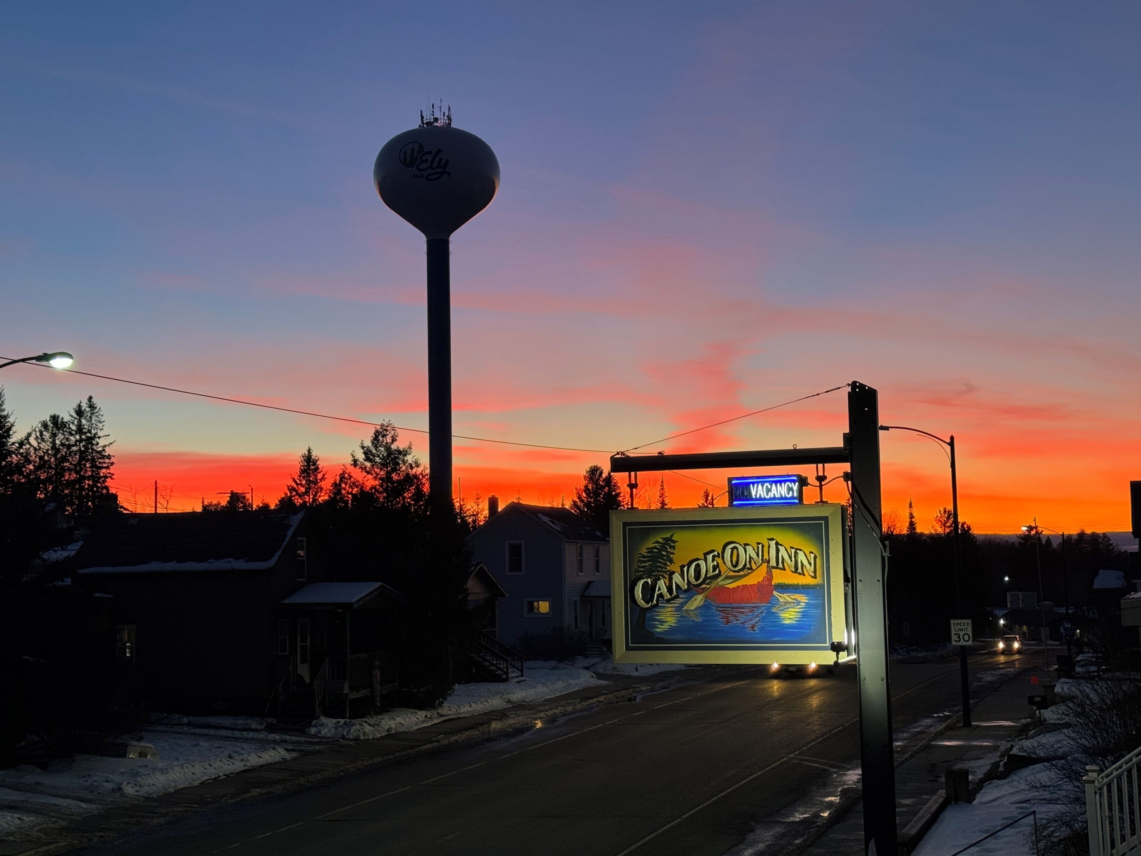 A hotel sign with a "VACANCY" sign above it, a speed limit sign, and a water tower in the background.