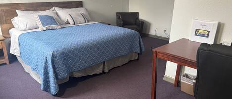 A hotel room with a bed, a desk, a chair, a lamp, and a welcome sign.