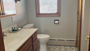 Un baño con inodoro blanco, un lavabo con tocador de madera, una ventana con persianas y suelo de baldosas.