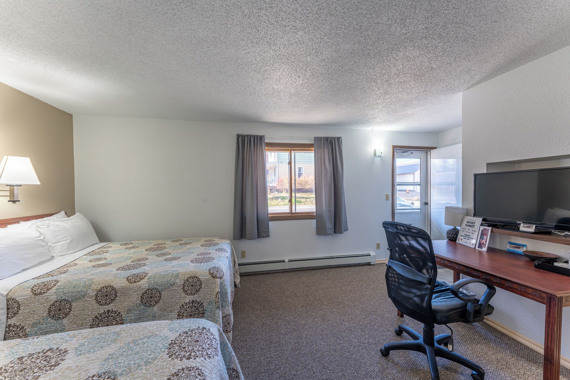 A hotel room with two beds, a desk with a computer and TV, and a view of the outside.
