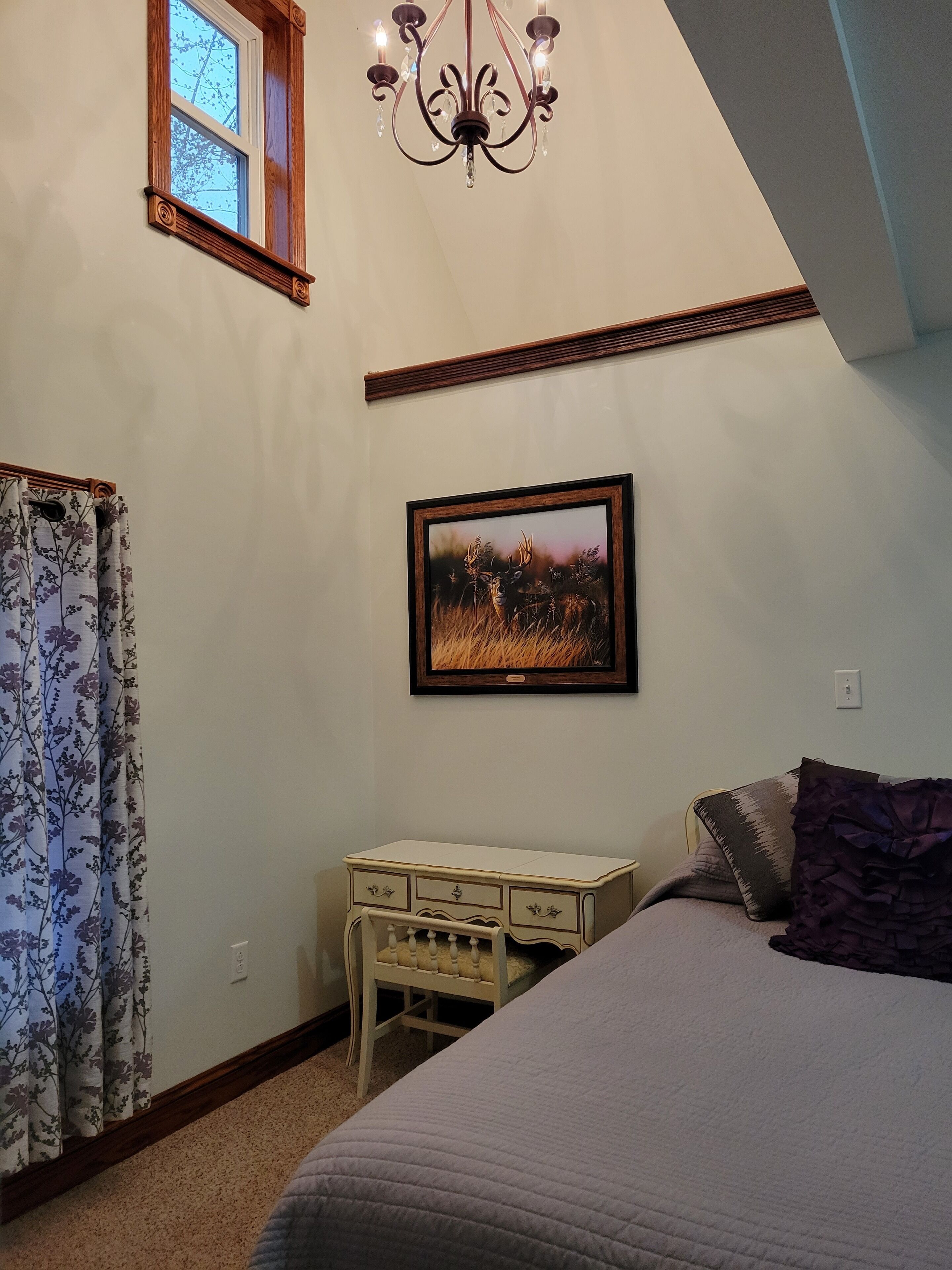 A bedroom with a bed, a desk, a chandelier, a window, and a framed picture on the wall.
