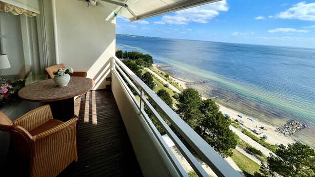 Un balcón con mesa y sillas, con vista a una playa de aguas cristalinas y una costa distante.