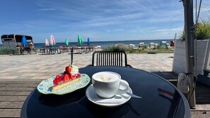 Een tafel met een stuk taart en een kop koffie, met uitzicht op een strand met stoelen en parasols.