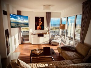 A modern living room with a large TV, a sofa, a coffee table, and a balcony with a view. - Moin Mehrblick – Penthouse near the Beach with Terrace and Wi-Fi (Schillig)