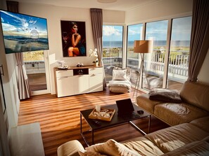 A modern living room with a sofa, a coffee table, a TV, and a balcony with a view of the sea. - Moin Mehrblick – Penthouse near the Beach with Terrace and Wi-Fi (Schillig)