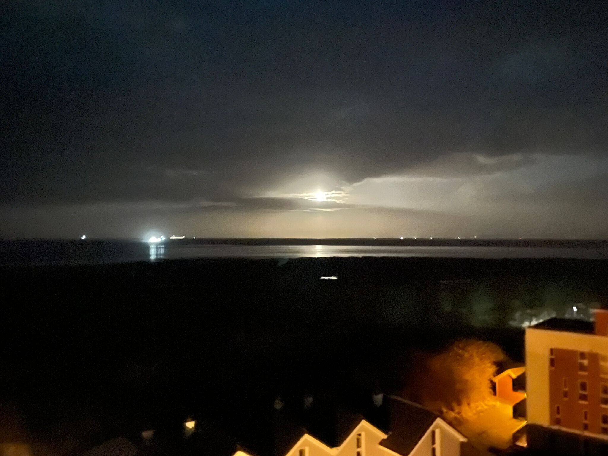 Ein Nachtbild einer Küstenlandschaft mit weiter Sicht auf ein Gewässer, einem bewölkten Himmel und einigen Gebäuden im Vordergrund.