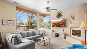A living room with a gray sofa, a blue armchair, a coffee table, a fireplace, and large windows offering a view of trees and a desert landscape. - Luxury Sedona Retreat • Hot Tub & Fire Pit (Sedona)