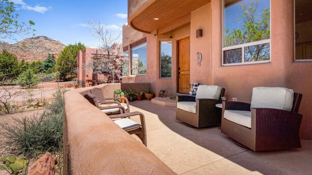 A modern outdoor patio with wicker furniture, a wooden door, and large windows overlooking a mountainous landscape. - Luxury Sedona Retreat • Hot Tub & Fire Pit (Sedona)