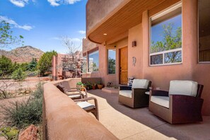 A modern outdoor patio with wicker furniture, a wooden door, and large windows overlooking a mountainous landscape. - Luxury Sedona Retreat • Hot Tub & Fire Pit (Sedona)