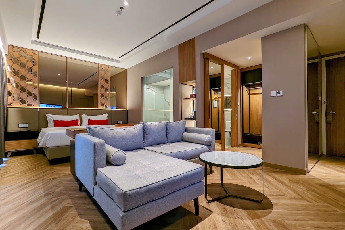 Une chambre d’hôtel moderne avec un canapé d’angle gris, une petite table ronde, un grand miroir et un plancher en bois.