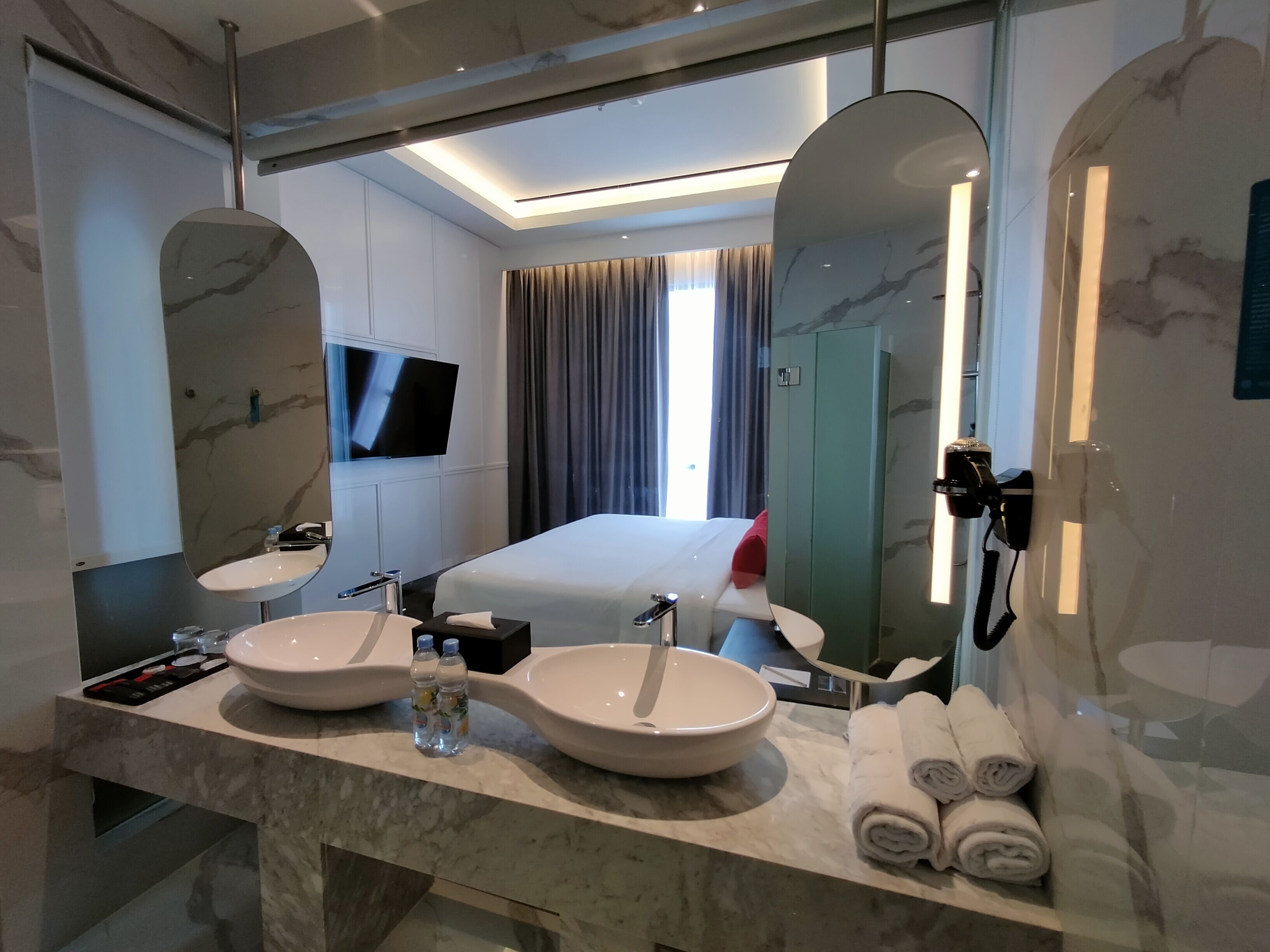 A modern hotel bathroom with two large sinks, a mirror, a flat-screen TV, and a bed with a view of the city.