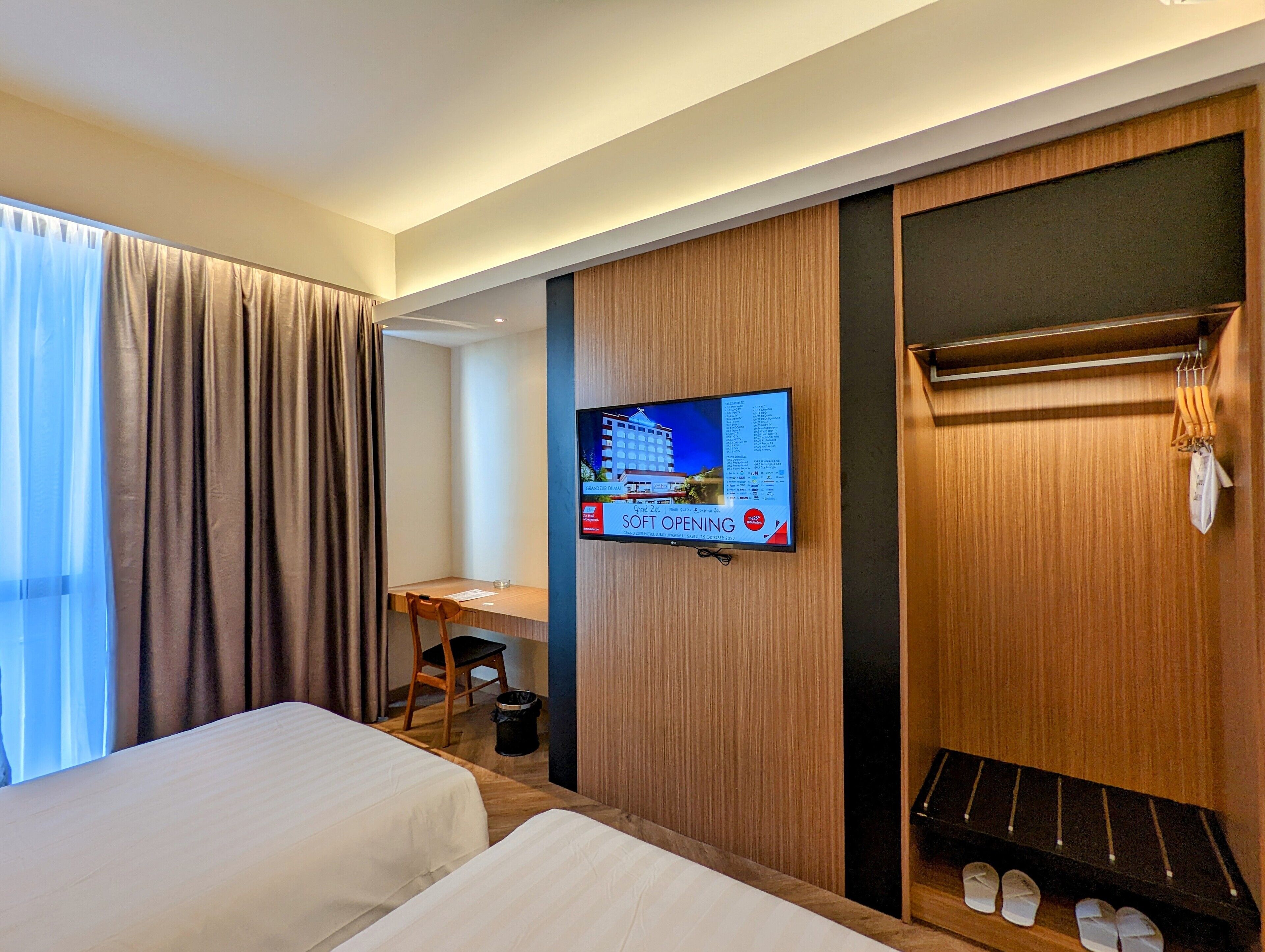 A hotel room with two beds, a desk, a television mounted on the wall, and a shoe rack.