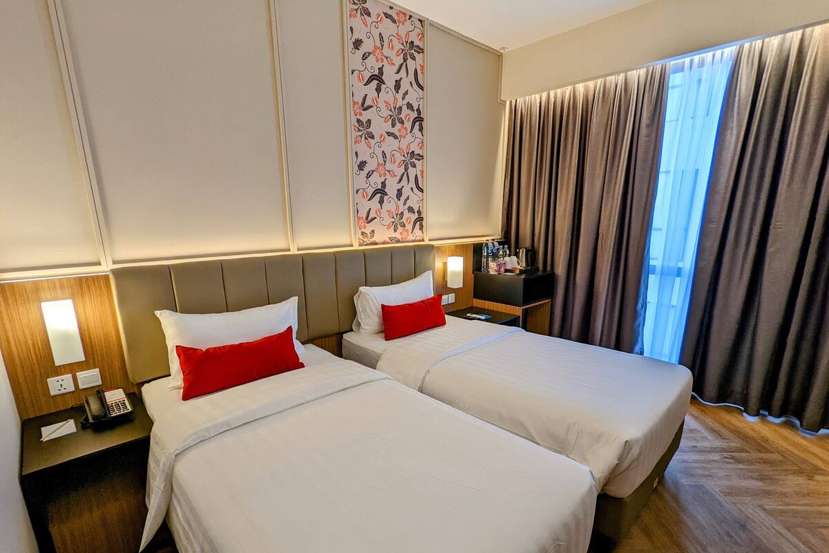 Une chambre d’hôtel avec deux lits, une table de chevet avec un téléphone, une lampe fixée au mur et une fenêtre avec des rideaux.