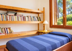Een kamer met een bed, houten planken met boeken, een klein tafeltje met een lamp en een raam met luiken.