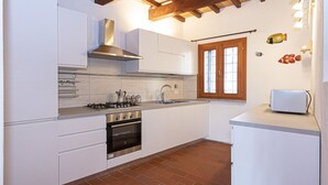 Een keuken met witte kasten, een gasfornuis, een oven, een spoelbak en een raam met houten kozijnen.