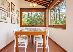A dining area with a glass-top table and white chairs, large windows with a view of trees, and framed pictures on the wall.