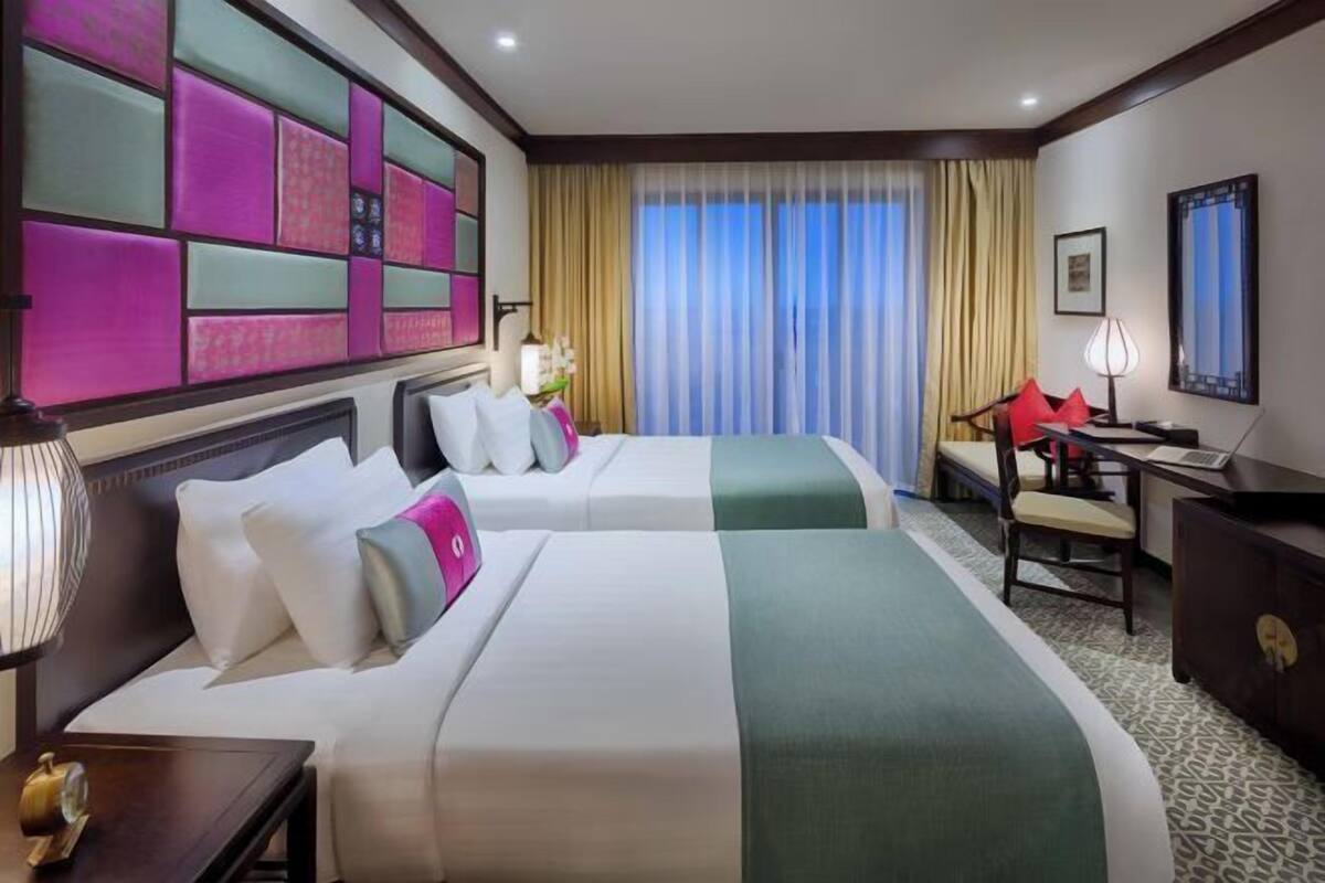A hotel room with two beds, a desk, a chair, a lamp, and a large window with curtains.