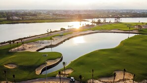 Un campo de golf con búnkeres de arena, un obstáculo acuático y un atardecer de fondo.