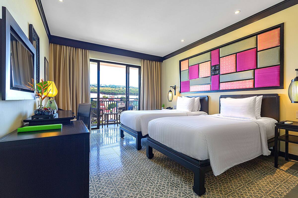 A hotel room with two beds, a desk, a chair, a balcony with a view, and a colorful wall art piece.
