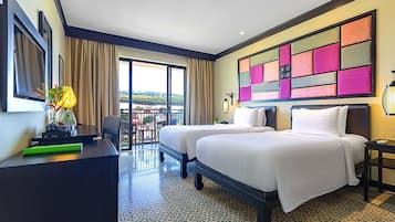 Une chambre d’hôtel avec deux lits, un bureau, une chaise, un balcon avec vue et une œuvre d’art murale aux couleurs vives.