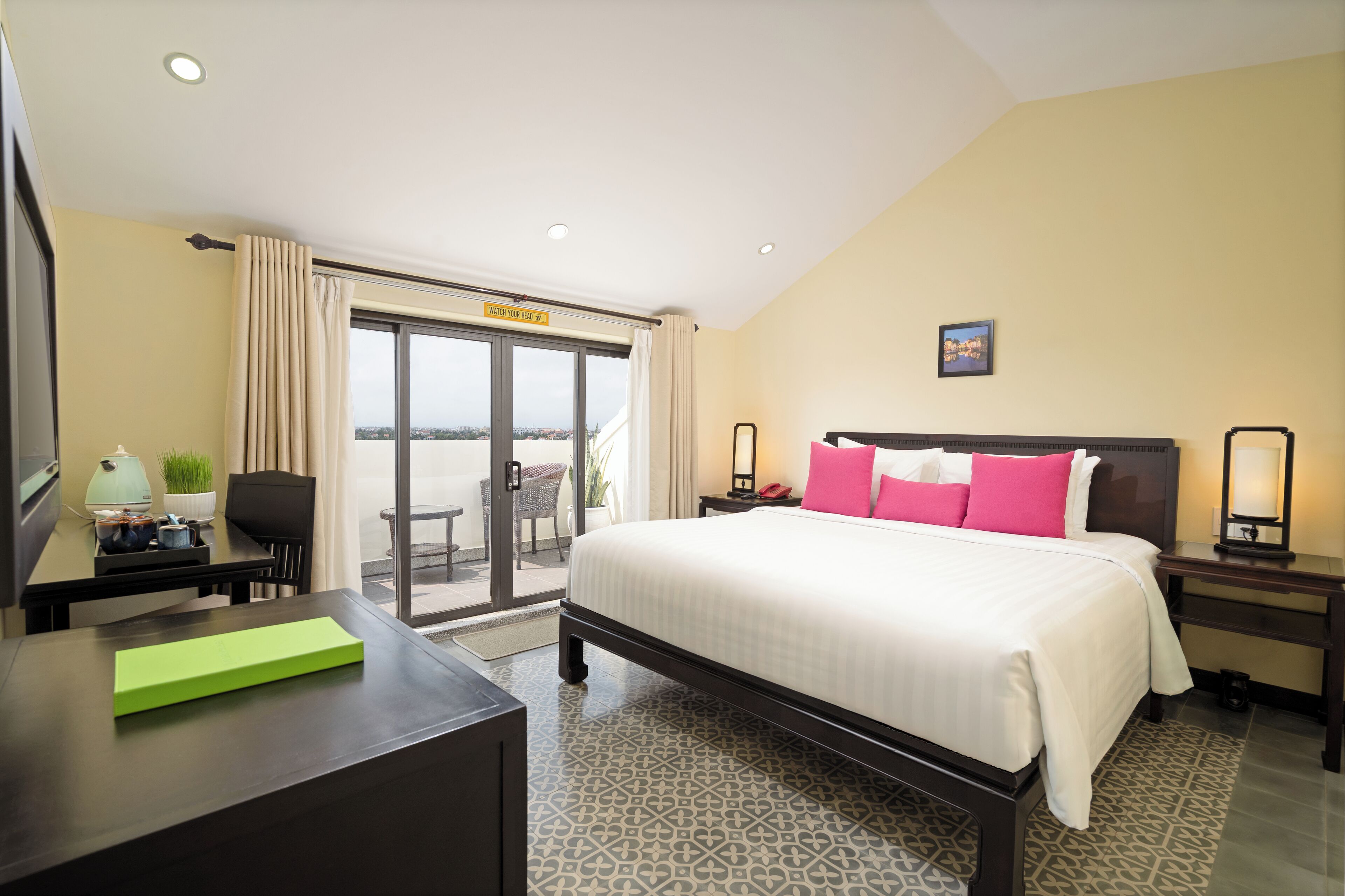 A hotel room with a large bed, a desk, a chair, a small table, a nightstand, a lamp, a picture on the wall, and a view of the outside through a sliding door.