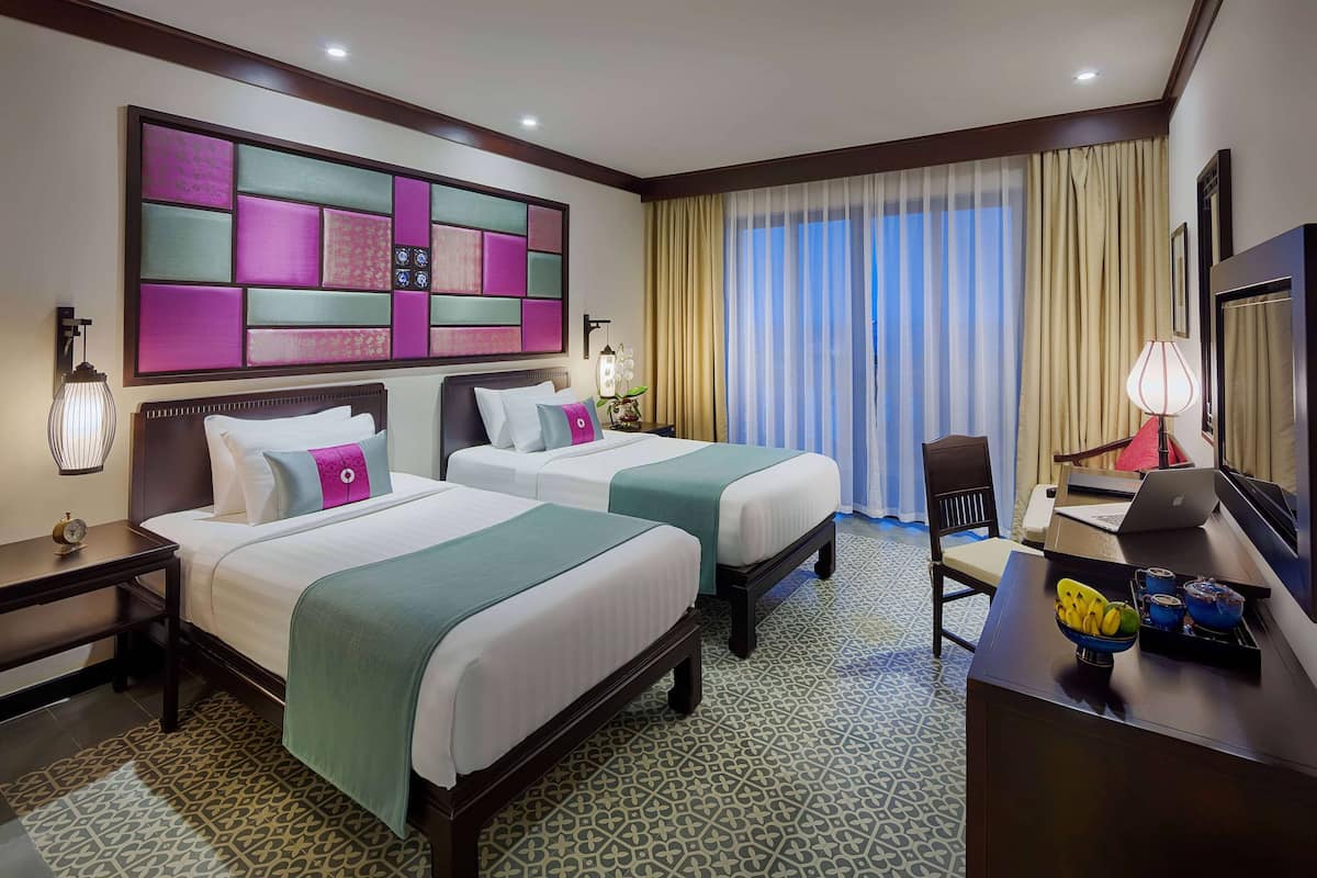 A hotel room with two beds, a desk with a laptop, a chair, a decorative wall, and a large window with curtains.