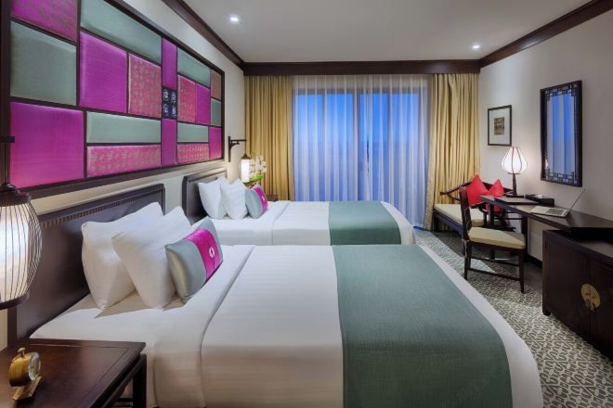 Quarto de hotel com duas camas, uma escrivaninha, uma cadeira, uma luminária e uma janela grande com cortinas.