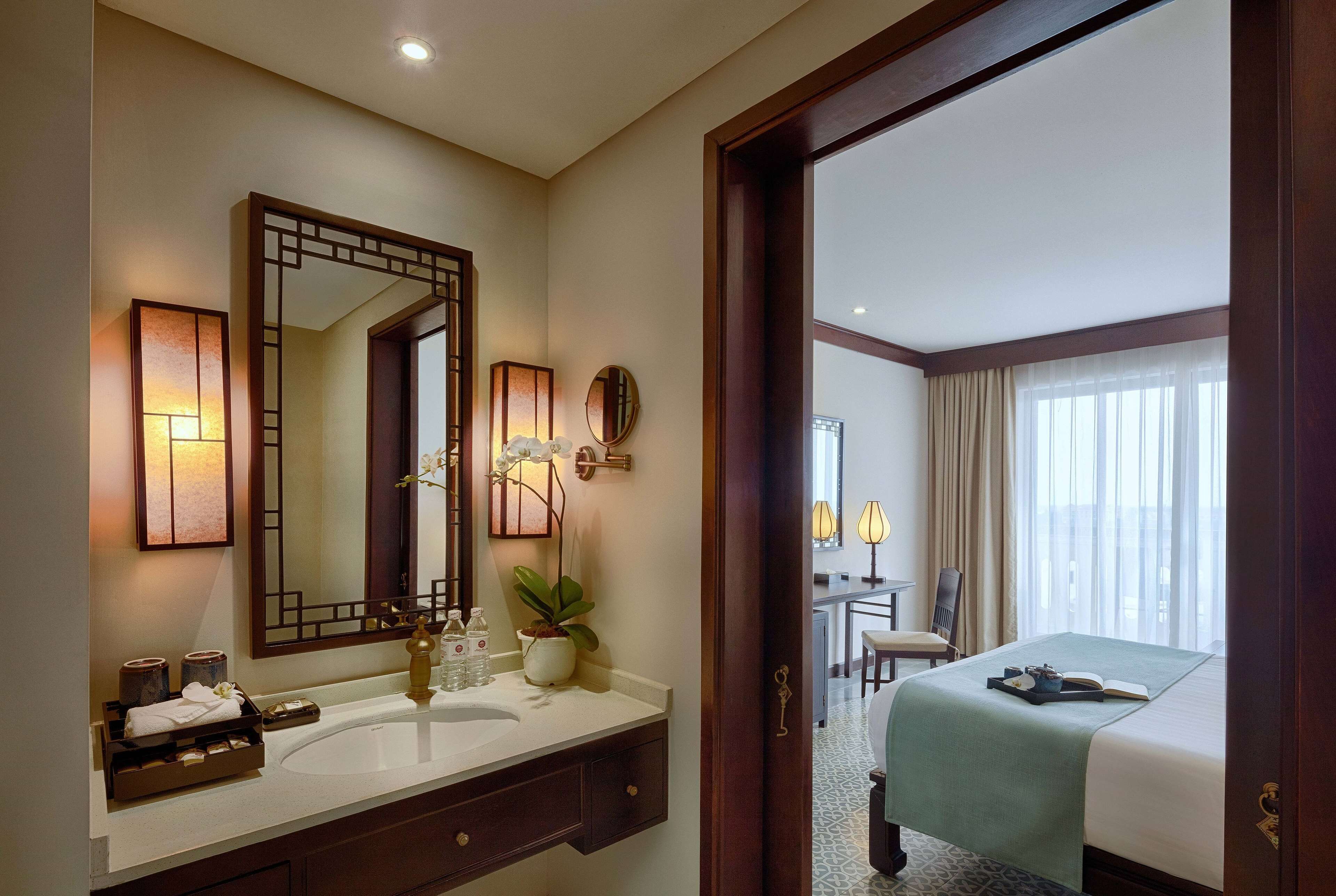 A hotel room with a large mirror, a sink, a bedside table with a lamp, a bed, and a view of the outside through a window.