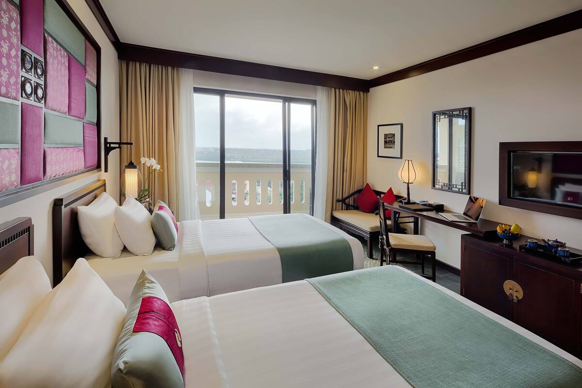 A hotel room with two beds, a desk, a chair, a TV, and a balcony with a view.