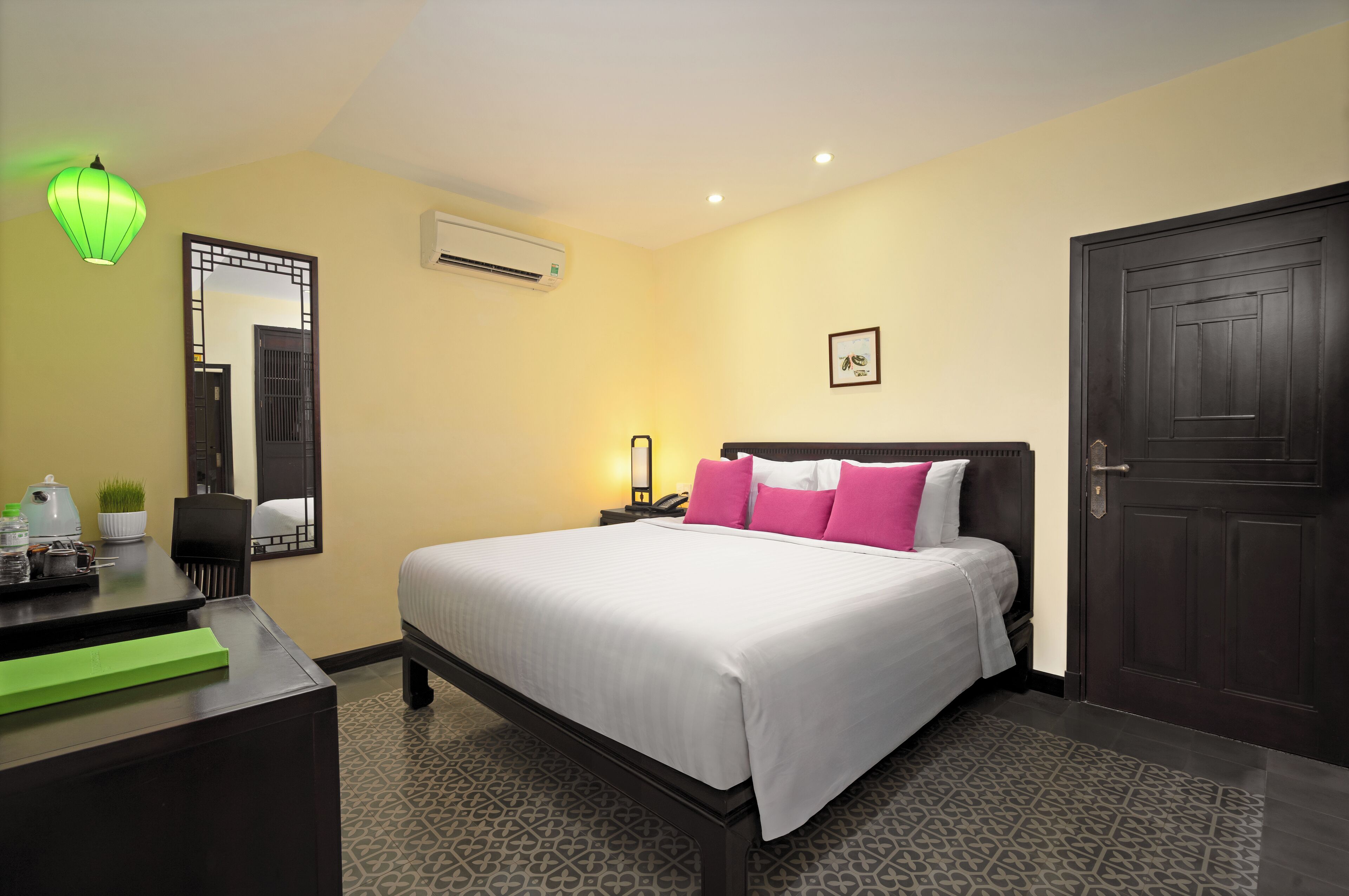 A hotel room with a bed, bedside table, lamp, air conditioning unit, and a door.