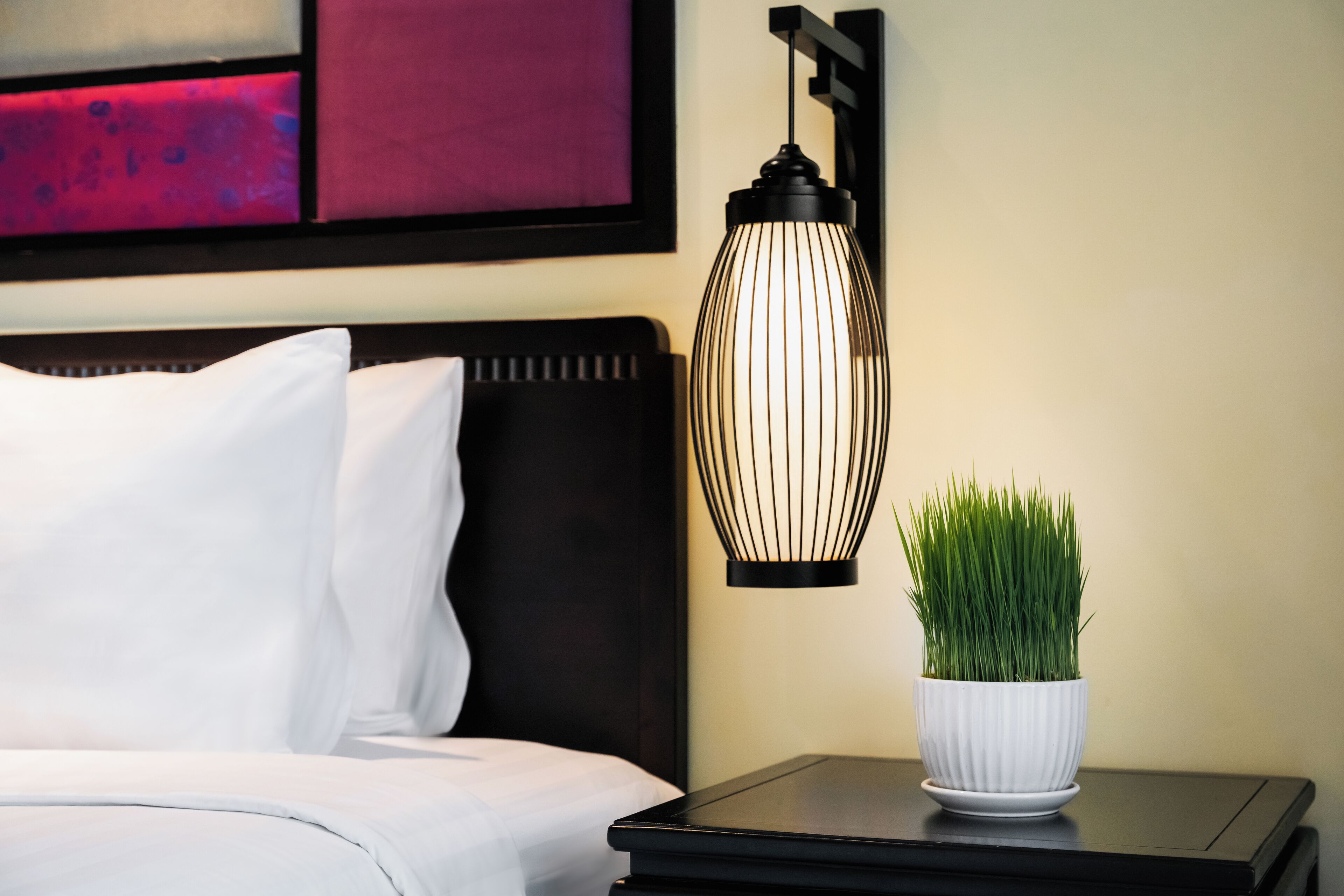 A neatly made bed with white linens, a black nightstand with a white pot of green plants, and a modern wall-mounted light fixture.