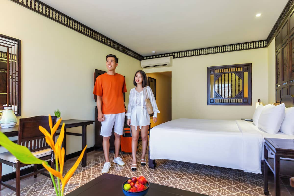 A hotel room with a bed, a desk, a chair, a small table with a bowl of fruit, and a decorative plant.