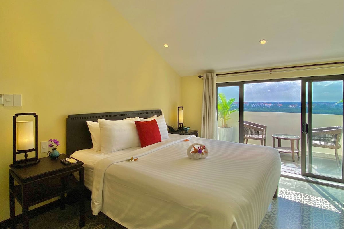 A hotel room with a large bed, a nightstand with a lamp, a vase with flowers, and a view of the outside balcony.