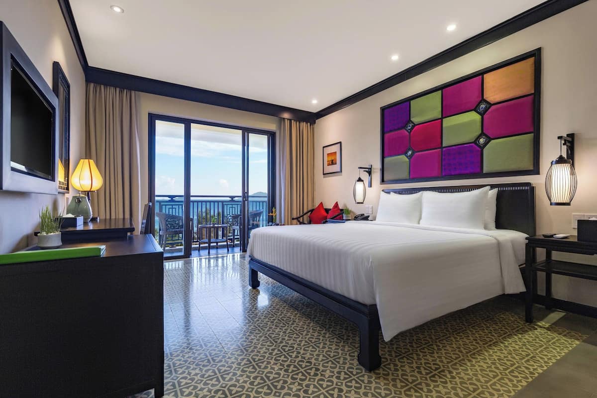 A hotel room with a large bed, a desk, a TV, a balcony with a view, and a colorful wall panel.