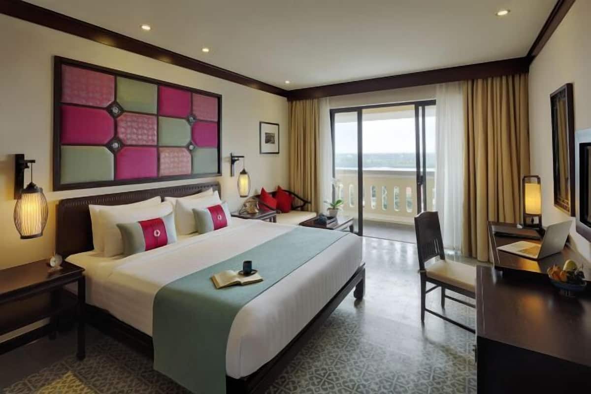 A hotel room with a large bed, a desk with a laptop, a chair, a balcony with curtains, and a decorative wall piece.