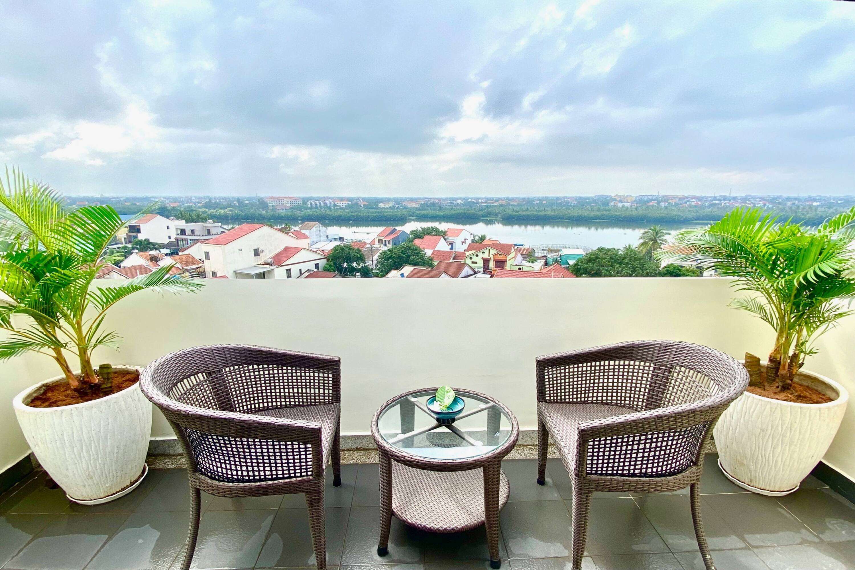 A balcony with two wicker chairs and a small table, overlooking a cityscape with buildings and a body of water.