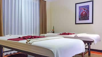 A room with two beds, each covered with white linens and a red blanket. There is a framed picture on the wall and a window with curtains.
