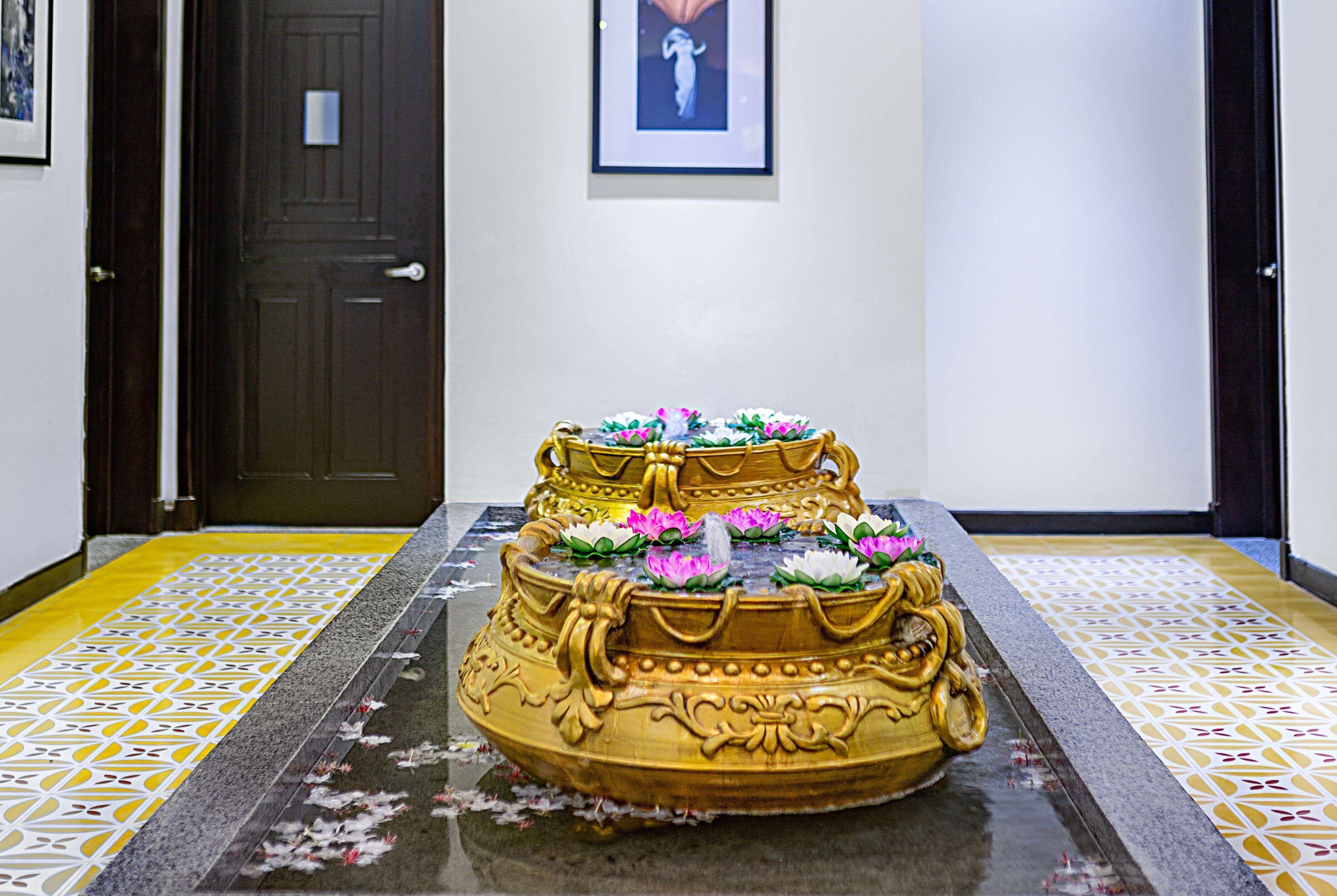 A three-tiered golden fountain with water lilies, set on a patterned floor with a door and framed artwork in the background.