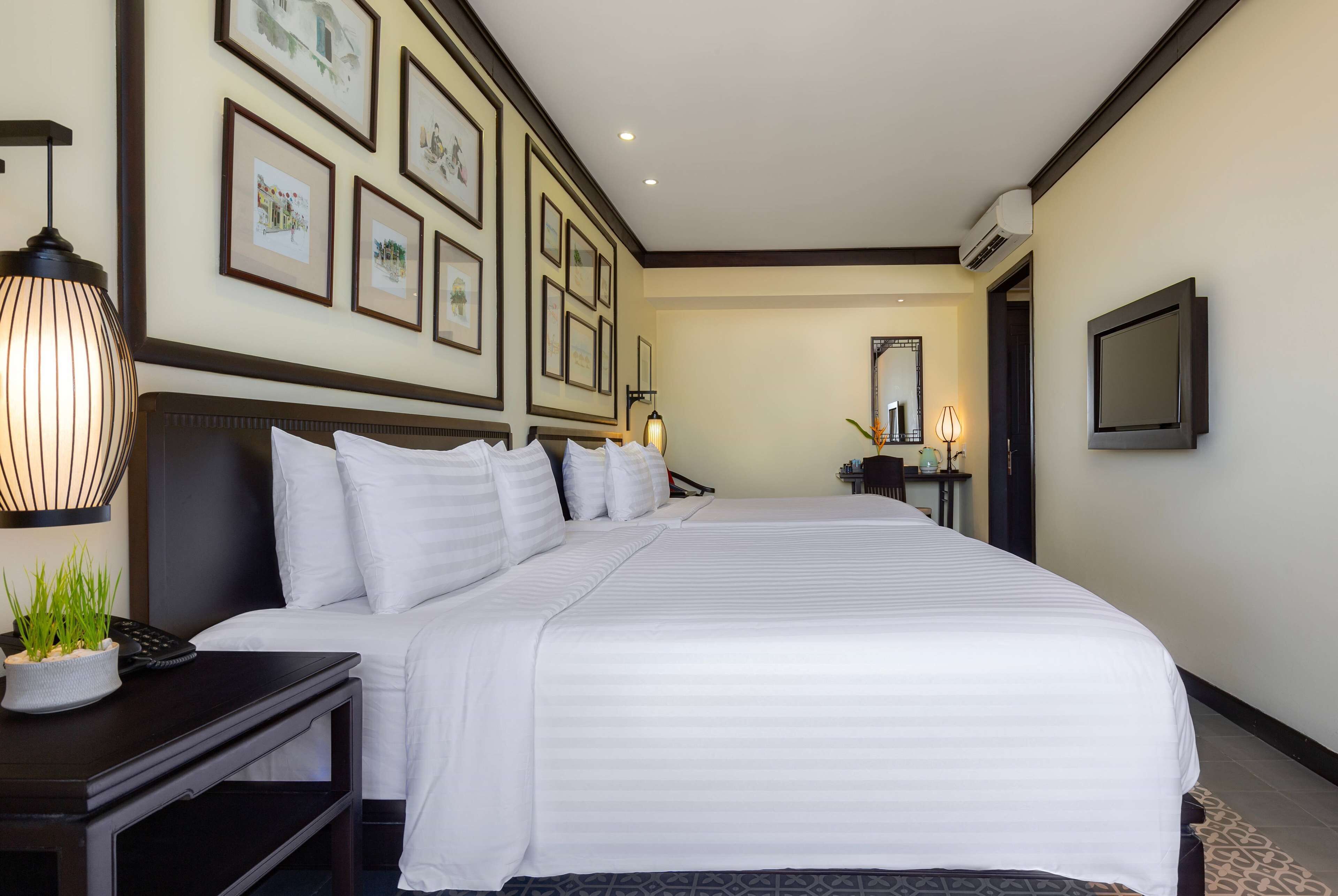 A hotel room with a large bed, a bedside table with a lamp, a small plant, and framed pictures on the wall.